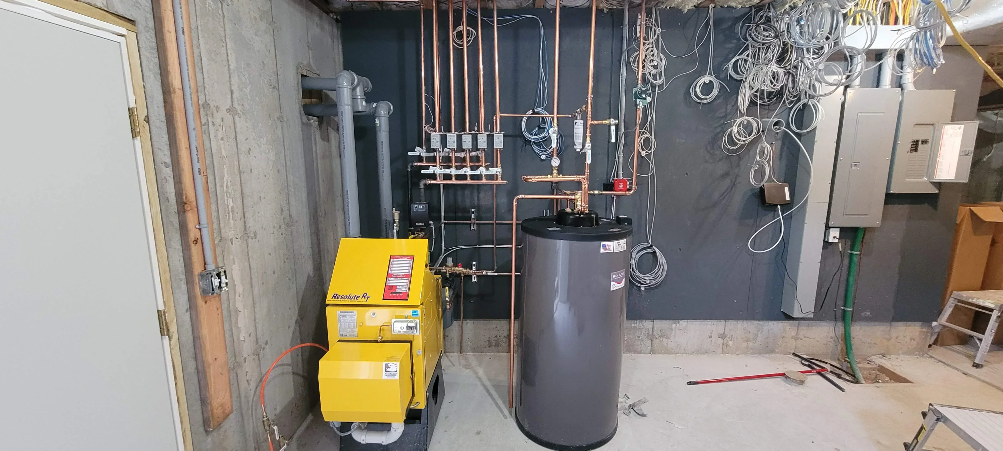 Yellow boiler unit with gray storage tank, copper piping, and radiant heating distribution manifolds in mechanical room