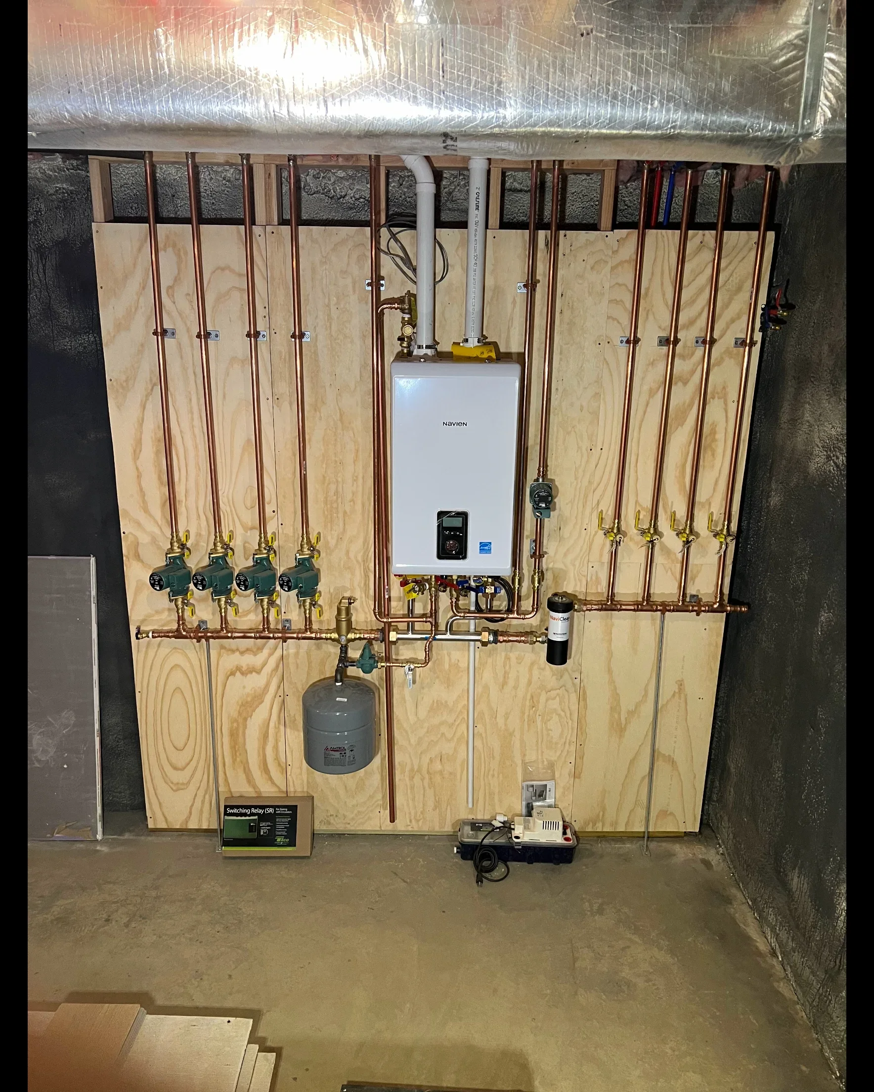 Tankless water heater with copper piping, radiant heat manifold, expansion tank, and multi-zone valve controls on plywood backing